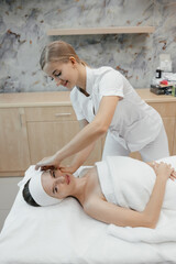 Shot of cosmetologist making face massage for rejuvenation to woman while lying on a stretcher in the spa center. Relaxed young female is resting on couch while cosmetologist massaging her face