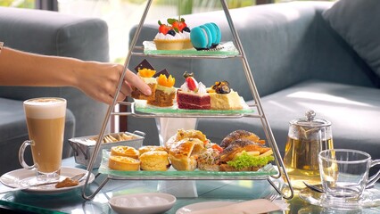 Woman hand take slice of cake from a towering display of afternoon tea - obrazy, fototapety, plakaty