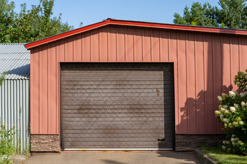 Modern red garage with driveway and roller door. Wide parking gate with shadows of trees and...