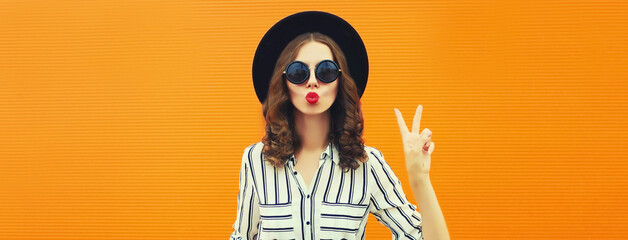 Summer portrait of beautiful young woman model blowing her lips sending sweet air kiss wearing black round hat, striped shirt on orange background