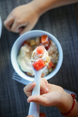 Little girl eating muesli with yoghurt and fruits with white plastic spoon