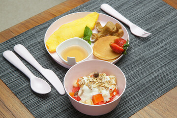 Kids menu - omlet, pancakes with maple syrup, muesli with yoghurt and glass of fresh squuzed orange juice served on the restaurant table with plastic kids friendly eating utensil.