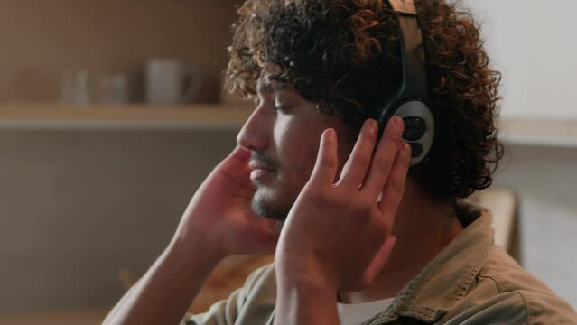 Close up Indian guy with curly hair wearing headphones dancing to song in kitchen. Cheerful relaxed Arabian man male dancer homeowner in earphones listen to music dance moving active to audio at home