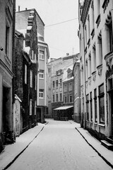 Black and white vertical photo of narrow old town street and vintage buildings covered in snow in winter. Old town Riga, Latvia