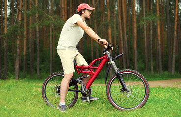 Naklejka premium Sport, leisure and healthy lifestyle concept - young man sitting on a bicycle in the forest, male cyclist in profile on grass against the trees