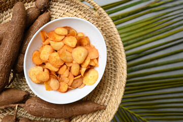 Organic yuca or cassava chips in bowl and tapioca root on palm leaves. Indonesia popular hot spicy snacks. Top view. Copy space.