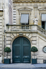 Paris ancient stone building with wooden carved double doors, stone staircase, French windows with striped awning canvas blinds, statue, rich stucco fretwork, downspout and potted plants in front