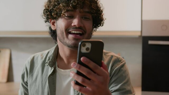 Indian man with curly hair holding mobile phone Arabian guy talking to webcam has online virtual meeting greeting hello talk to smartphone video call conference chat with friends at kitchen at home