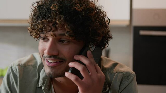 Cheerful Indian young man with curly hair talking phone in home kitchen. Happy friendly Hispanic guy talk with friends by smartphone Arabian male speaking smiling laughing pleasant mobile conversation