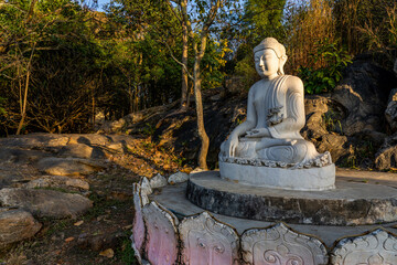 White meditating Buddha statue sits on the rock pile among forest trees in the afternoon time at India. Great religious architecture. Hight quality photo