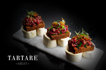 Side view of portion of raw beef tartare served on rye bread toasts with micro green on top. Minced meat dish on veal bones and marble plate. Dish isolated on black background. Ready menu banner