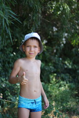 A white European schoolboy in swimming trunks and a hat shows a thumbs-up gesture against the...