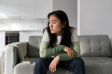 Sad lonely young Asian woman having depression symptoms, sitting on sofa at home looking into distance, upset unhappy female worrying about money or family problems. Mental disorders, 