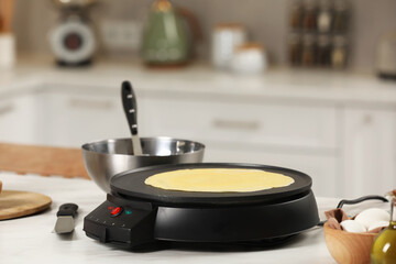 Cooking delicious crepe on electric pancake maker on white marble table in kitchen, space for text - obrazy, fototapety, plakaty