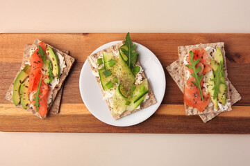 Fresh crunchy crispbreads with cream cheese, cucumber, green onion, salmon and arugula on beige table, top view