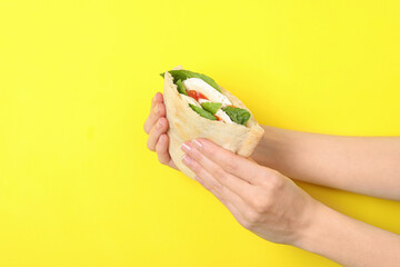 Woman holding delicious pita sandwich with mozzarella, tomatoes and basil on yellow background,...
