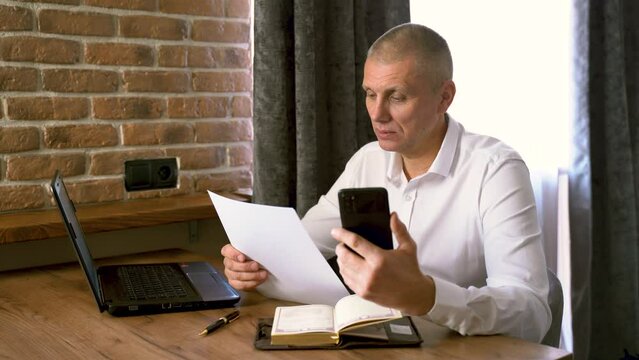 A businessman holds a phone and documents in his hands, talks via video link, checks information. Financial manager, company.