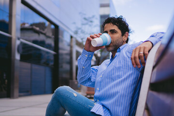 Middle-aged Latin man sitting on a city bench drinking coffee from a thermos cup. Copy space.