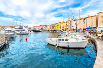 Sailboats and luxury yachts line the colorful marina filled with shops and seaside cafes in the town of Saint-Tropez, France, a popular resort town on the Mediterranean Cote d'Azur, French Riviera - obrazy, fototapety, plakaty