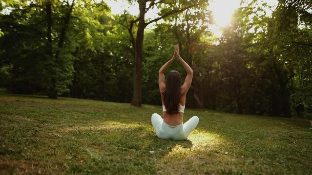 Back view of unrecognizable slender young woman sitting on yoga mat in lotus position and raising hands up outside in city park. Rear view of female practicing yoga outdoors in sunny day.