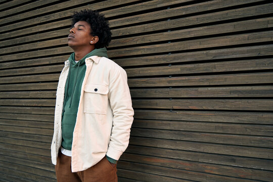 Cool young hipster gen z African American teen standing at wooden wall. Sensitive vulnerable ethnic rebel teenage boy breathing air with eyes closed feeling free or sad in city outdoors.