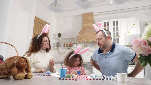 Beautiful family having Easter breakfast in their cozy kitchen. Portrait of little girl enjoying painting colorful eggs with her mother and father. High quality 4k footage