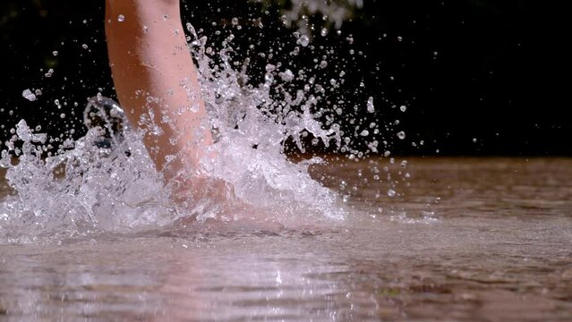 SLOW MOTION, CLOSE UP: Female bare feet walking in shallow wild mountain river . Drops of water splash while she is crossing the pristine river barefoot. Pleasant refreshment in hot sunny summer days.