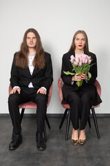 Obraz premium Elegant couple wearing black suits. A woman looking into the camera. A man holding a tulip and looking at it. Grey background. Place for your text.