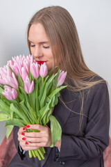 Elegant beautiful busines woman in black suit. Woman holding a bouquet of pink tulips and smells them. Grey background.