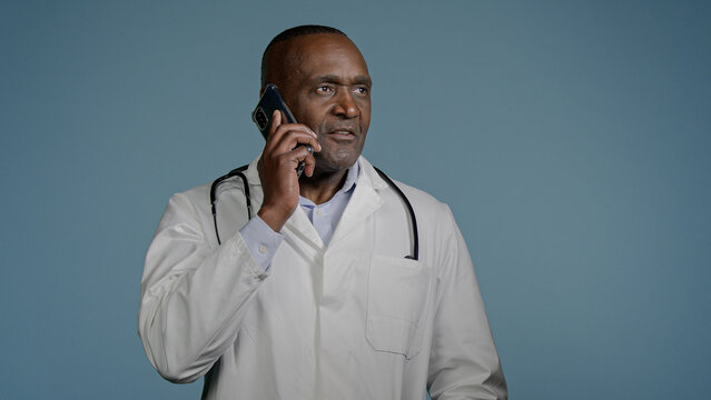 Mature African American Man Doctor Talking On Phone With Patient In Blue Background Studio Male Therapist In Medical Gown Consult Client Using Smartphone Remote Medical Assistance Healthcare Concept