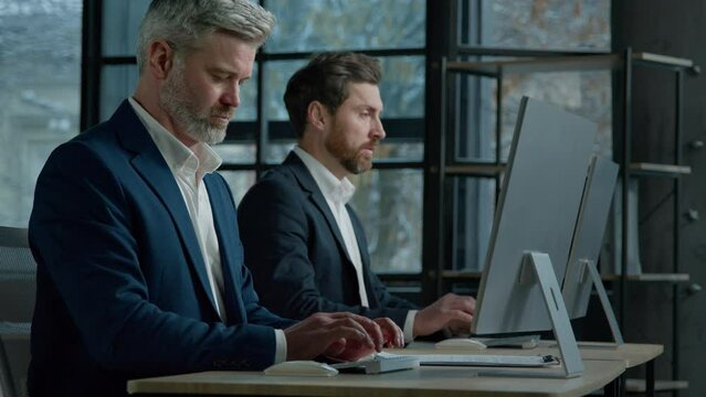 Focused professional managers working with computers on project sit in row at desk in office caucasian adult business men businessmen corporate employees colleagues work on Internet typing on computer
