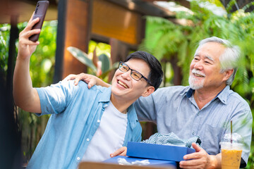 Happy Asian man using mobile phone taking selfie with elderly father during celebrating birthday party together at restaurant on summer vacation. Family relationship older people health care concept.