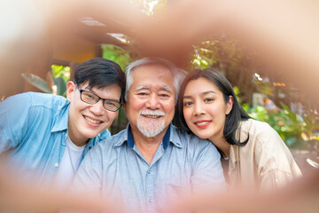 Happy Asian couple using mobile phone taking selfie with elderly father during having lunch eating food together at restaurant on summer vacation. Family relationship older people health care concept.