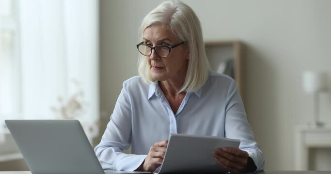 Concentrated middle-aged businesswoman working with documents sits at desk with laptop, look through utility bills, check bills, read agreement details, prepare report, analyzes data, manage finances