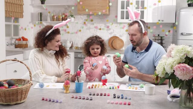 Father and mother showing their cute little toddler how to eat traditional colorful eggs. Portrait of a baby girl with curly hair painting and decorating Easter table. High quality 4k footage
