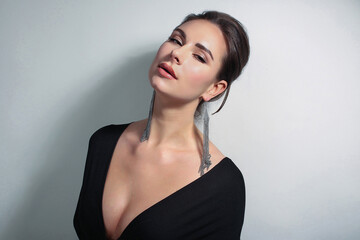 Portrait of a beautiful fashionista brunette girl, with long silver earrings, in black with a neckline on a light white background in the studio room