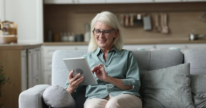 Happy senior woman sit on comfy couch in cozy room, use digital tablet for entertainment, browse social networks, watch funny video, laughing at joke, chat with friends, shopping online, surf website
