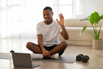 Cheerful african sportsman waving at laptop screen, home interior