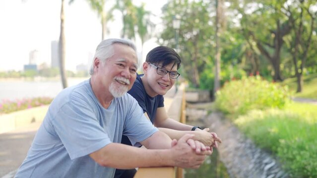 Asian adult son and elderly father looking beautiful nature during jogging exercise together at park. Retired man enjoy outdoor sport workout. Family relationship and older people health care concept.