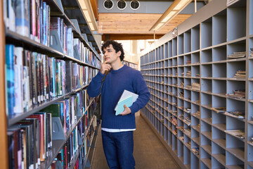 Pensive male student, thoughtfully looking at bookshelves, looking for books in the library campus