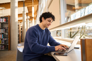Smart Hispanic male student sitting at desk and using laptop preparing diploma project in the...