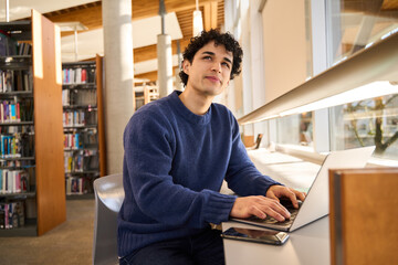 Pensive young Hispanic male student online working on laptop in a modern innovation library campus