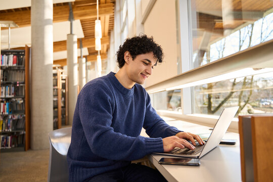 Smart Hispanic male student sitting at desk and using laptop preparing diploma project in the library campus