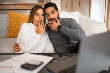 Shocked scared millennial european female hugs arab husband with beard, watch on laptop in living room