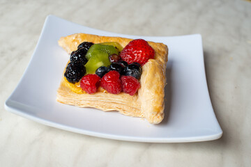 A square fruit filled French puff pastry on a white square plate. The cream and custard filled dessert have strawberries, kiwi, and blackberries on top with a sugar glaze. The pastry is flaky.	
