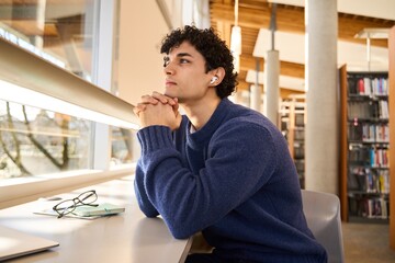 Hispanic student attentively listens to a broadcasted online lesson on earphones, in library campus - obrazy, fototapety, plakaty