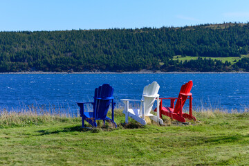 Three Adirondack chairs one red, blue, and white sit on the edge of the ocean on a sunny summer's...