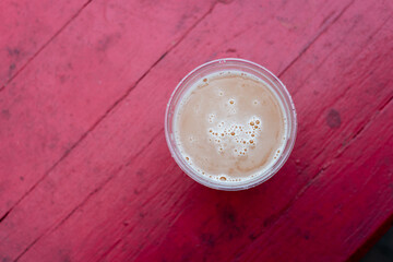 A top-down view of a clear glass of Belgian beer. The creamy carbonated draft head has froth foam and air bubbles of gas rising. The brew ale is on a vibrant red wooden plank table at a microbrewery. 