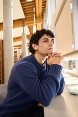 Pensive Hispanic male student sitting at desk in the modern innovation university library campus