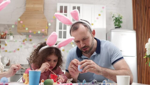 Adorable toddler with curly hair wearing bunny ears and painting with her parents. Portrait of little girl helping dad and mom preparing for Easter holiday. High quality 4k footage